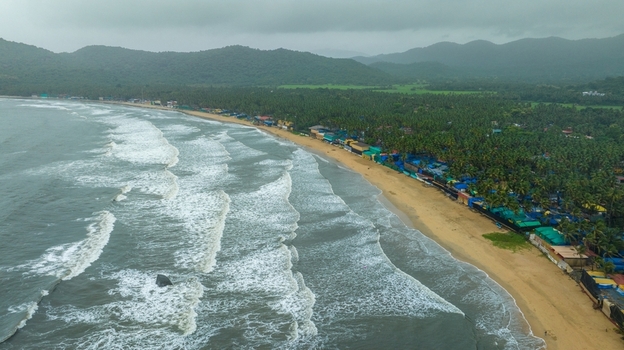 Aerial view of Palolem Beach with waves crashing on the sandy shore lush palm trees and green hills in the background under a cloudy sky in Canacona Goa India.