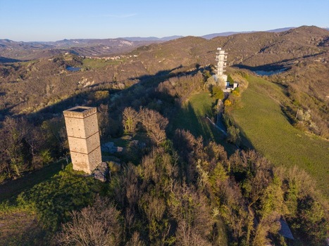 Aerial view of Television Tower Tortona and the historic stone tower on a wooded hilltop surrounded by rolling green hills and dense forest in Tortona Piedmont Italy.