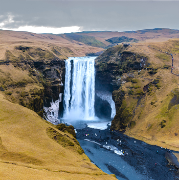 Aerial view of Skógafoss waterfall cascading powerfully into the dark river below framed by golden hills under a cloudy sky Skógafoss Rangárþing eystra Iceland. Aerial view of Skógafoss waterfall cascading powerfully into the dark river below framed by golden hills under a cloudy sky Skógafoss Rangárþing eystra Iceland.