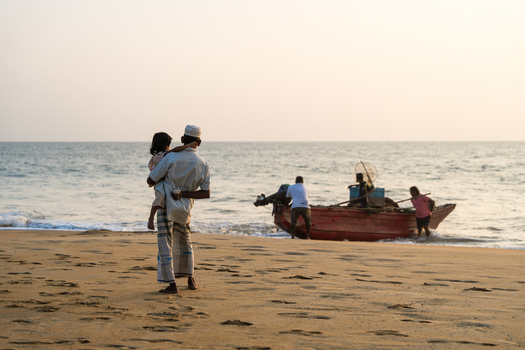 Sri Lanka 23 January 2026 View of a man holding a child on a sandy beach while fishermen prepare a traditional red boat in the ocean at sunset.