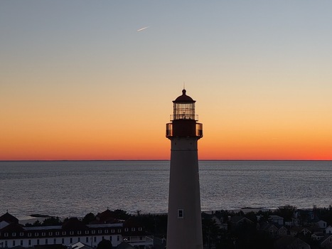 Aerial view of Cape May Lighthouse silhouetted against a vibrant orange sunset sky over the Atlantic Ocean with a distant plane contrail Cape May New Jersey United States.