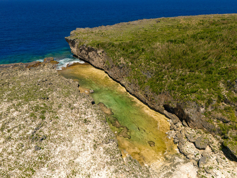 Aerial view of Chigit Beach featuring rugged limestone cliffs turquoise shallow waters and lush green vegetation in Northern Tinian Tinian Northern Mariana Islands. Aerial view of Chigit Beach featuring rugged limestone cliffs turquoise shallow waters and lush green vegetation in Northern Tinian Tinian Northern Mariana Islands.