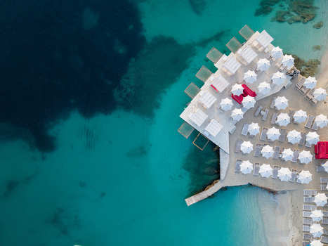 Aerial view of a luxury beach club with white umbrellas and wooden decks over turquoise sea water in Ksamil Vlore County Albania. Aerial view of a luxury beach club with white umbrellas and wooden decks over turquoise sea water in Ksamil Vlore County Albania.