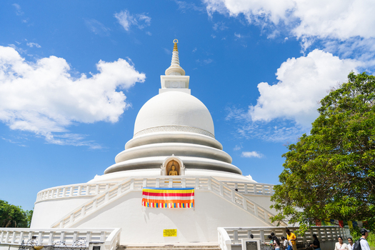 View of the white Japanese Peace Pagoda Rumassala with visitors and lush greenery under a blue sky in Unawatuna Southern Province Sri Lanka. View of the white Japanese Peace Pagoda Rumassala with visitors and lush greenery under a blue sky in Unawatuna Southern Province Sri Lanka.