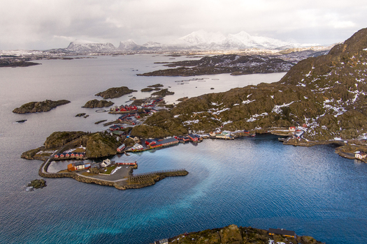 Aerial view of Lofoten featuring traditional red wooden houses nestled among rocky terrain and deep blue fjord waters with snow capped mountains in the distance Nordland Norway.