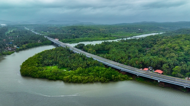 Aerial view of a long bridge spanning over the Galgibaga River surrounded by lush green palm trees and dense mangrove forests under a cloudy sky in Canacona Goa India.