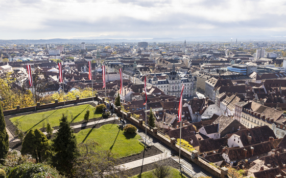 View of red and white flags fluttering above the green landscape the city sprawling with buildings and rooftops under a cloudy sky Graz Styria Austria. View of red and white flags fluttering above the green landscape the city sprawling with buildings and rooftops under a cloudy sky Graz Styria Austria.