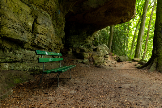 View of a green bench on the Mullerthal Trail path under a sandstone rock formation in a forest in Berdorf Echternach Luxembourg. View of a green bench on the Mullerthal Trail path under a sandstone rock formation in a forest in Berdorf Echternach Luxembourg.