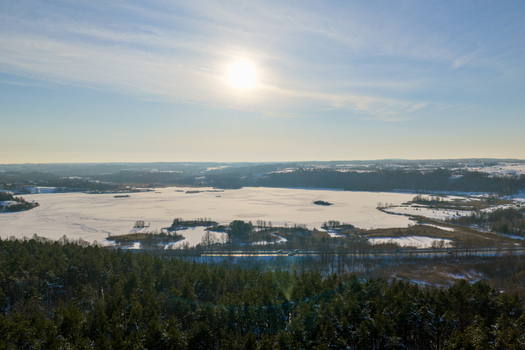 Aerial view of a frozen lake surrounded by snow covered fields and dense evergreen forests under a bright winter sun in Lithuania. Aerial view of a frozen lake surrounded by snow covered fields and dense evergreen forests under a bright winter sun in Lithuania.