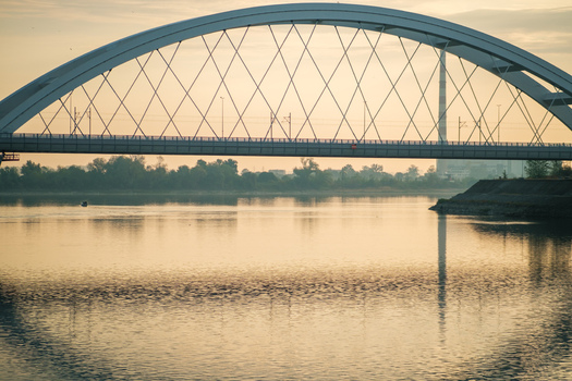 View of Zezelj Bridge spanning the Danube River at sunset with calm water reflections and a distant industrial chimney in Novi Sad Vojvodina Serbia. View of Zezelj Bridge spanning the Danube River at sunset with calm water reflections and a distant industrial chimney in Novi Sad Vojvodina Serbia.