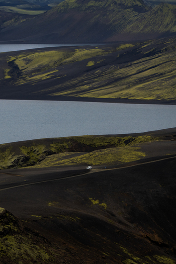 View of a lone white vehicle traverses a dark volcanic landscape bisecting the scene between the stark black earth and the tranquil blue waters Skaftárhreppur Iceland. View of a lone white vehicle traverses a dark volcanic landscape bisecting the scene between the stark black earth and the tranquil blue waters Skaftárhreppur Iceland.
