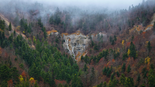 Aerial view of a misty verdant mountainside cascading into a delicate waterfall framed by autumn kissed trees under a cloudy sky Gex Auvergne Rhône Alpes France. Aerial view of a misty verdant mountainside cascading into a delicate waterfall framed by autumn kissed trees under a cloudy sky Gex Auvergne Rhône Alpes France.