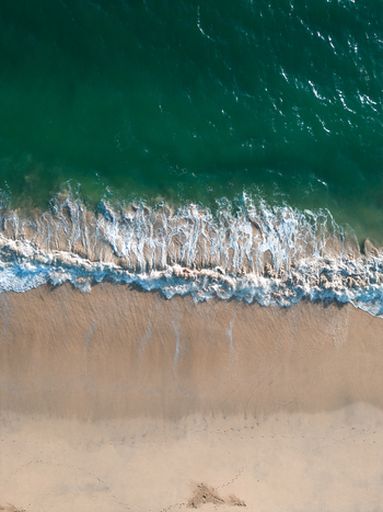 Aerial view of turquoise ocean waves crashing onto a sandy shore with white sea foam and footprints in the sand Carbis Bay Cornwall United Kingdom.