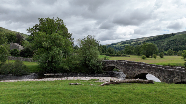 View of an ancient stone arch bridge crossing a flowing river surrounded by lush green meadows and rolling hills under a cloudy sky in Hawes England United Kingdom. View of an ancient stone arch bridge crossing a flowing river surrounded by lush green meadows and rolling hills under a cloudy sky in Hawes England United Kingdom.