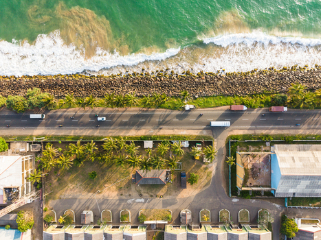 Aerial view of coastal road with vehicles driving past palm trees and a rocky breakwater by the turquoise ocean in Galle Southern Province Sri Lanka.