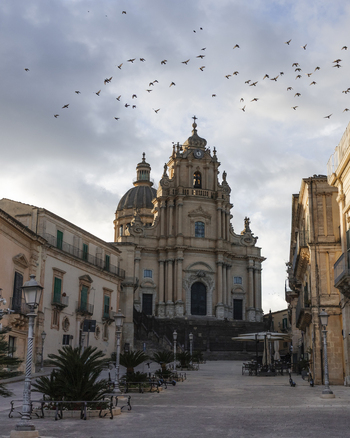 View of the baroque San Giorgio Cathedral with a flock of birds flying over the historic square and traditional buildings in Ragusa Sicily Italy.