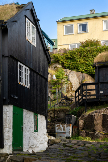View of a traditional black wooden house with a green turf roof and a bright green door set against a rocky hillside with a yellow house in the background in the Faroe Islands. View of a traditional black wooden house with a green turf roof and a bright green door set against a rocky hillside with a yellow house in the background in the Faroe Islands.