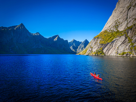 Aerial view of a red kayak with two people paddling in deep blue water surrounded by steep rocky mountains in Hamnoy Nordland Norway.