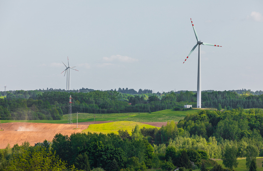View of wind turbines standing in a lush green rural landscape with rolling fields dense forests and power lines under a clear sky in Goldap Warmian Masurian Voiv Poland.