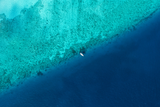 Aerial view of a small white boat floating in the transition between shallow turquoise coral reef waters and the deep blue ocean in Vashafaru Haa Alif Atoll Maldives. Aerial view of a small white boat floating in the transition between shallow turquoise coral reef waters and the deep blue ocean in Vashafaru Haa Alif Atoll Maldives.