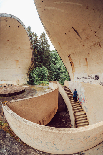 View of the brutalist concrete monument of the Korcanica Memorial Zone with a person walking on stairs surrounded by trees in Sanski Most Federation of Bosnia Bosnia and Herzegovina. View of the brutalist concrete monument of the Korcanica Memorial Zone with a person walking on stairs surrounded by trees in Sanski Most Federation of Bosnia Bosnia and Herzegovina.