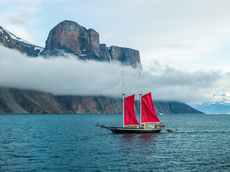 Aerial view of a sailboat with vibrant red sails navigating the deep blue waters of a fjord near the towering cliffs of Storoen in Avannaata Greenland. Aerial view of a sailboat with vibrant red sails navigating the deep blue waters of a fjord near the towering cliffs of Storoen in Avannaata Greenland.