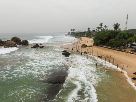 Aerial view of traditional stilt fishermen on the sandy beach with waves crashing against rocks and lush palm trees in Koggala Southern Province Sri Lanka. Aerial view of traditional stilt fishermen on the sandy beach with waves crashing against rocks and lush palm trees in Koggala Southern Province Sri Lanka.