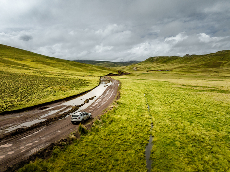 Aerial view of a car driving on a muddy unpaved road through a vast green hilly landscape under a cloudy sky in Canas Cuzco Peru. Aerial view of a car driving on a muddy unpaved road through a vast green hilly landscape under a cloudy sky in Canas Cuzco Peru.
