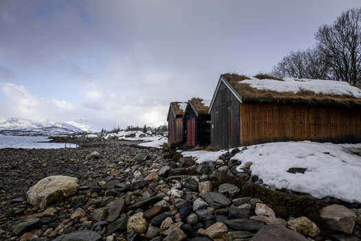 View of traditional wooden cabins with turf roofs on a snowy rocky shoreline under an overcast sky with mountains in the distance Kvaloya Troms Norway. View of traditional wooden cabins with turf roofs on a snowy rocky shoreline under an overcast sky with mountains in the distance Kvaloya Troms Norway.