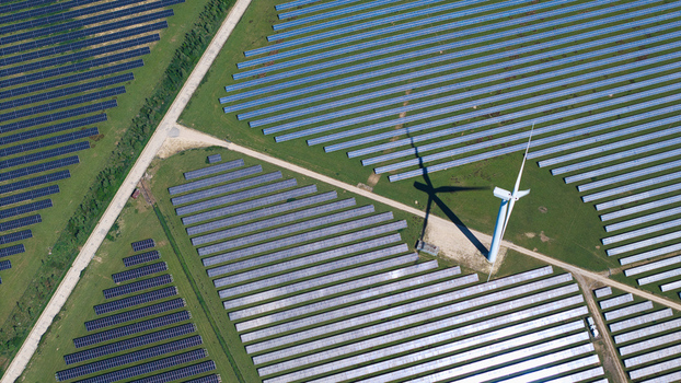 Aerial view of Chelveston Wind Farm featuring rows of solar panels and a large wind turbine casting a shadow over green fields in Wellingborough England United Kingdom. Aerial view of Chelveston Wind Farm featuring rows of solar panels and a large wind turbine casting a shadow over green fields in Wellingborough England United Kingdom.