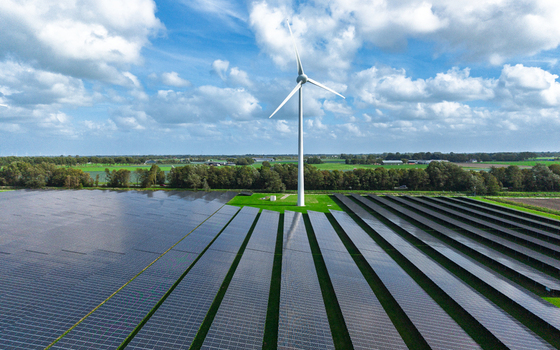 Aerial view of a large solar farm with a prominent wind turbine set against a backdrop of green fields and a blue sky with clouds in Warmenhuizen North Holland Netherlands.