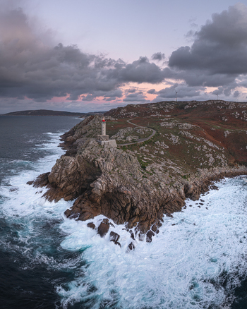 Aerial view of Faro de Punta Nariga on rocky cliffs with crashing white waves and wind turbines under a cloudy twilight sky Malpica A Coruna Spain. Aerial view of Faro de Punta Nariga on rocky cliffs with crashing white waves and wind turbines under a cloudy twilight sky Malpica A Coruna Spain.