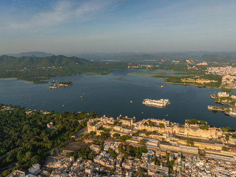 Aerial view of the historic City Palace complex on the banks of Lake Pichola featuring white architecture lush trees and calm lake waters Udaipur Rajasthan India.