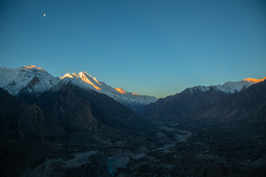 View of the snow capped Rakaposhi peaks glowing in golden light over the Hunza valley with a winding river under a clear blue sky with the moon Hunza Nagar Gilgit Pakistan. View of the snow capped Rakaposhi peaks glowing in golden light over the Hunza valley with a winding river under a clear blue sky with the moon Hunza Nagar Gilgit Pakistan.