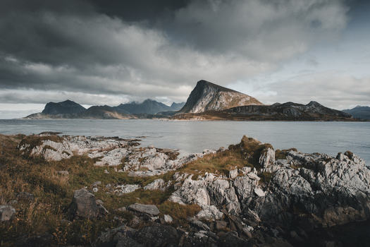 View of Myrland Beach with rugged rocky coastline calm sea and dramatic mountains under a cloudy sky in Napp Nordland Norway. View of Myrland Beach with rugged rocky coastline calm sea and dramatic mountains under a cloudy sky in Napp Nordland Norway.