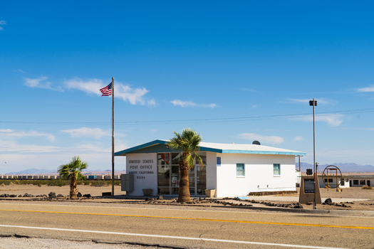 Amboy United States 06 April 2026 View of the United States Post Office building with an American flag and palm trees under a clear blue sky in the Mojave Desert.