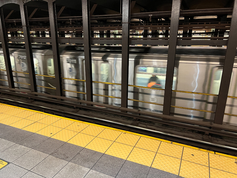 View of a New York City subway train moving past a station platform with yellow tactile paving and dark structural pillars New York New York United States. View of a New York City subway train moving past a station platform with yellow tactile paving and dark structural pillars New York New York United States.