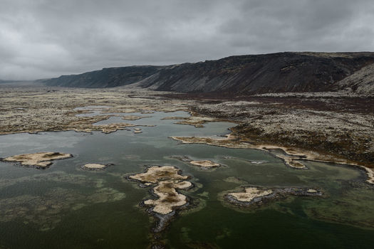 Aerial view of a volcanic landscape with moss covered lava fields scattered pools of water and a dark mountain ridge under a cloudy sky in Reykjavik Reykjavikurborg Iceland. Aerial view of a volcanic landscape with moss covered lava fields scattered pools of water and a dark mountain ridge under a cloudy sky in Reykjavik Reykjavikurborg Iceland.