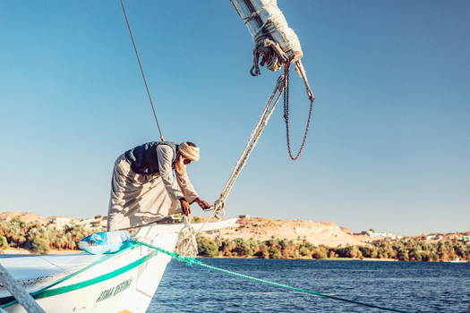 View of an Egyptian sailor in traditional clothing working on the bow of a felucca boat on the Nile river with desert hills in the background Aswan Aswan Governorate Egypt. View of an Egyptian sailor in traditional clothing working on the bow of a felucca boat on the Nile river with desert hills in the background Aswan Aswan Governorate Egypt.