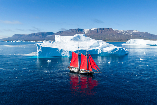 Aerial view of a sailboat with vibrant red sails navigating through deep blue waters surrounded by massive icebergs and distant snow capped mountains in Greenland. Aerial view of a sailboat with vibrant red sails navigating through deep blue waters surrounded by massive icebergs and distant snow capped mountains in Greenland.