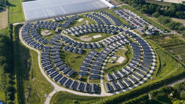 Aerial view of a holiday park with numerous small houses arranged in concentric circular patterns surrounded by lush green lawns and a large greenhouse complex in Venlo Limburg Netherlands. Aerial view of a holiday park with numerous small houses arranged in concentric circular patterns surrounded by lush green lawns and a large greenhouse complex in Venlo Limburg Netherlands.
