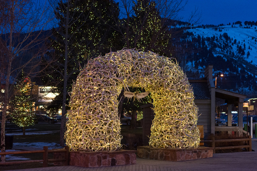 View of the illuminated elk antler arch in the town square at night with festive lights and snow capped mountains in the background in Teton County Wyoming United States.