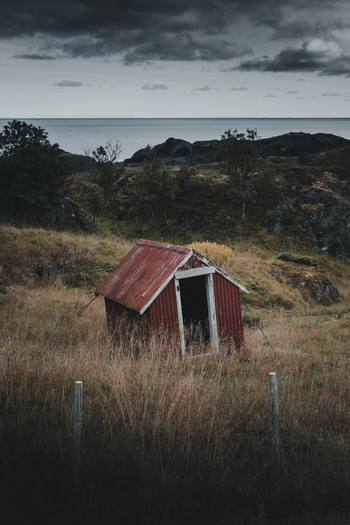View of the historic red wooden shed in Nesland surrounded by a grassy field and rocky coastal landscape in Flakstad Municipality Nordland Norway. View of the historic red wooden shed in Nesland surrounded by a grassy field and rocky coastal landscape in Flakstad Municipality Nordland Norway.