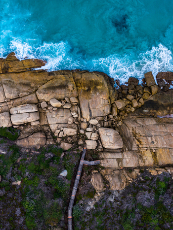Aerial view of turquoise waves crashing against granite cliffs and a wooden staircase winding through coastal vegetation in Torndirrup National Park Torndirrup Western Australia Australia.