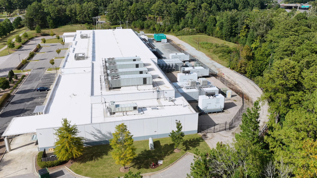 Aerial view of a large industrial building with a white roof and multiple HVAC units surrounded by a parking lot and dense forest in the United States. Aerial view of a large industrial building with a white roof and multiple HVAC units surrounded by a parking lot and dense forest in the United States.
