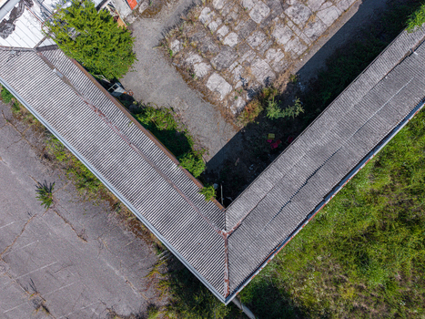 Aerial view of an L shaped industrial building with a corrugated metal roof surrounded by overgrown vegetation and asphalt in Casale Monferrato Piedmont Italy. Aerial view of an L shaped industrial building with a corrugated metal roof surrounded by overgrown vegetation and asphalt in Casale Monferrato Piedmont Italy.