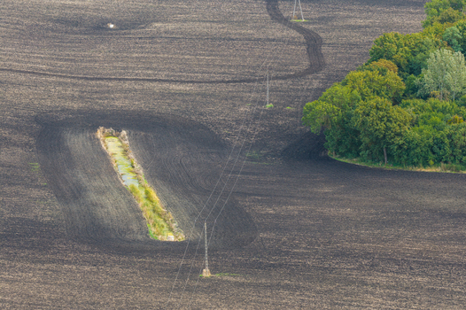 Aerial view of a dark plowed agricultural field with a narrow water filled ditch power lines crossing the landscape and a patch of green trees in Pomeranian Voivodeship Poland.