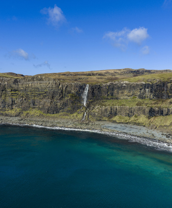 Aerial view of Talisker Bay Beach waterfall cascading down rugged cliffs into the turquoise sea under a clear blue sky Isle of Skye Scotland United Kingdom. Aerial view of Talisker Bay Beach waterfall cascading down rugged cliffs into the turquoise sea under a clear blue sky Isle of Skye Scotland United Kingdom.
