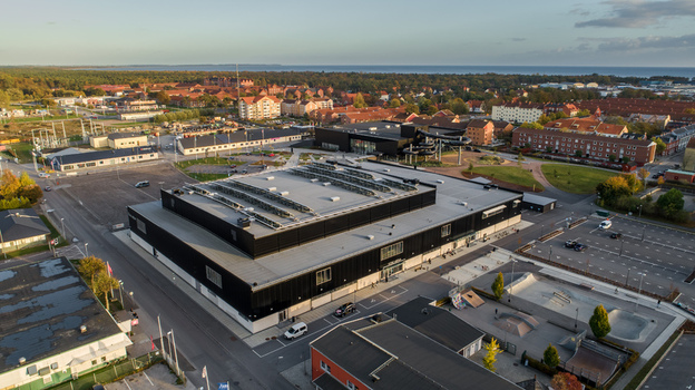 Aerial view of a modern sports arena with a large flat roof and solar panels adjacent to a skate park and urban residential buildings in Sweden. Aerial view of a modern sports arena with a large flat roof and solar panels adjacent to a skate park and urban residential buildings in Sweden.