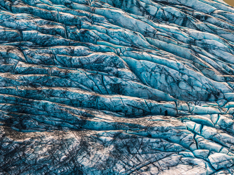 Aerial view of a glacier surface featuring deep blue crevasses and dark volcanic ash layers in Iceland. Aerial view of a glacier surface featuring deep blue crevasses and dark volcanic ash layers in Iceland.